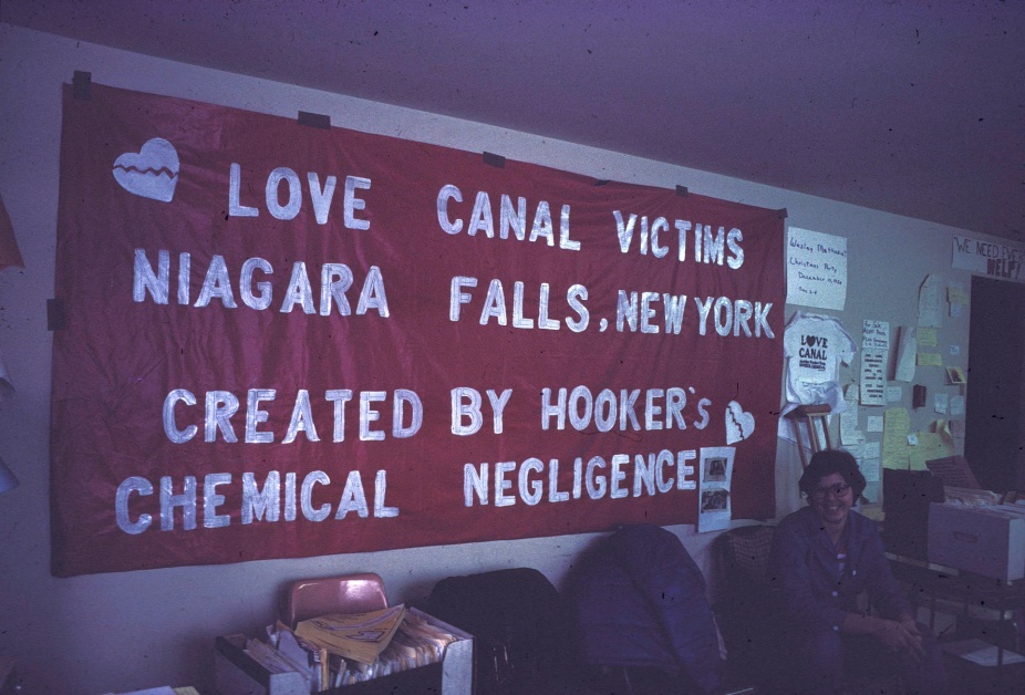 Debbie Cerrillo in the Love Canal Homeowners Association offices with banner reading, "Love Canal Victims, Niagara Falls, New York, Created by Hooker's chemical negligence," January 1981. 